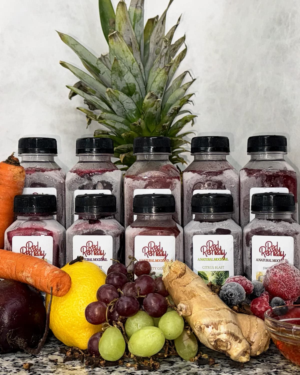 All five Red Remedy flavours, five 16 oz bottles stacked behind five 8 oz bottles, with a pineapple, carrots, a beet, a lemon, grapes, ginger, berries and honey
