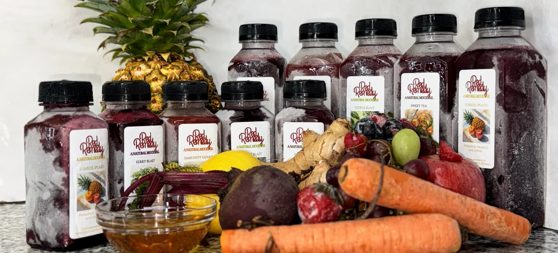 All five Red Remedy flavours in both sizes, five 8 oz bottles in front and five 16 oz bottles behind, with a pineapple, honey, a beet, a lemon, ginger, berries, grapes, carrots and an apple